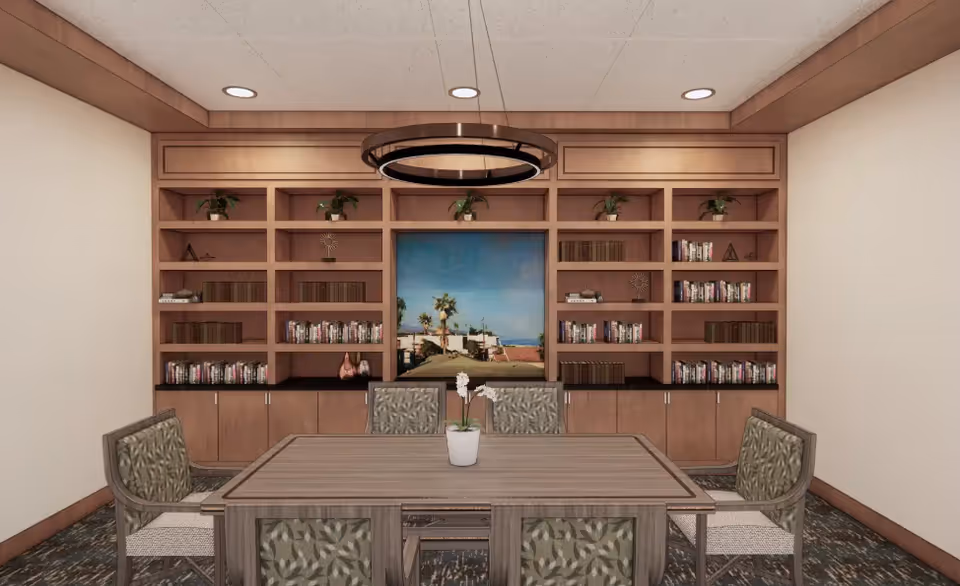 A small meeting or reading room with a wooden table surrounded by six patterned chairs. Behind the table is a large built-in wooden bookshelf filled with books and decorative plants. A framed picture of a beach scene with palm trees is centered on the bookshelf. The ceiling has recessed lighting and a modern circular hanging light fixture above the table.