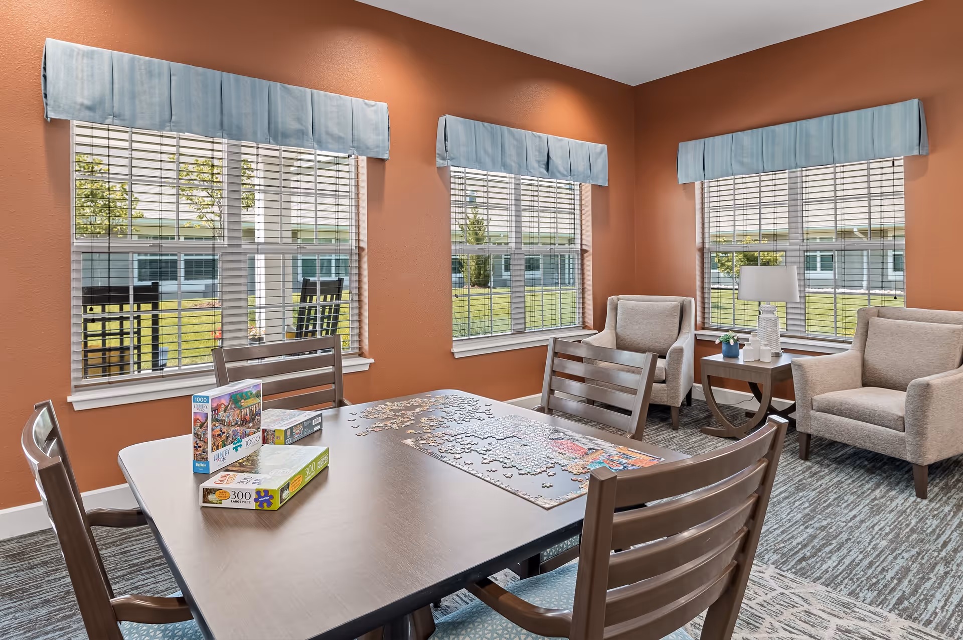 A cozy activity room with a wooden table and four chairs, featuring an incomplete jigsaw puzzle and puzzle boxes on the table. The room has three large windows with blue valances, letting in natural light and showing a green outdoor area. There are two beige armchairs and a small wooden side table with a lamp and a small plant in the corner.