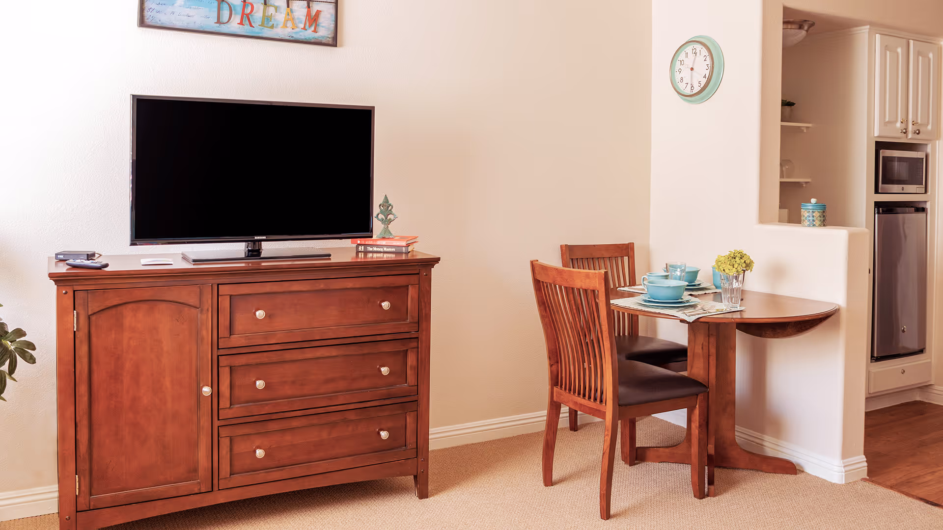 A cozy living area with a flat-screen TV on a wooden cabinet and a small dining table set for two beside a kitchen pass-through.
