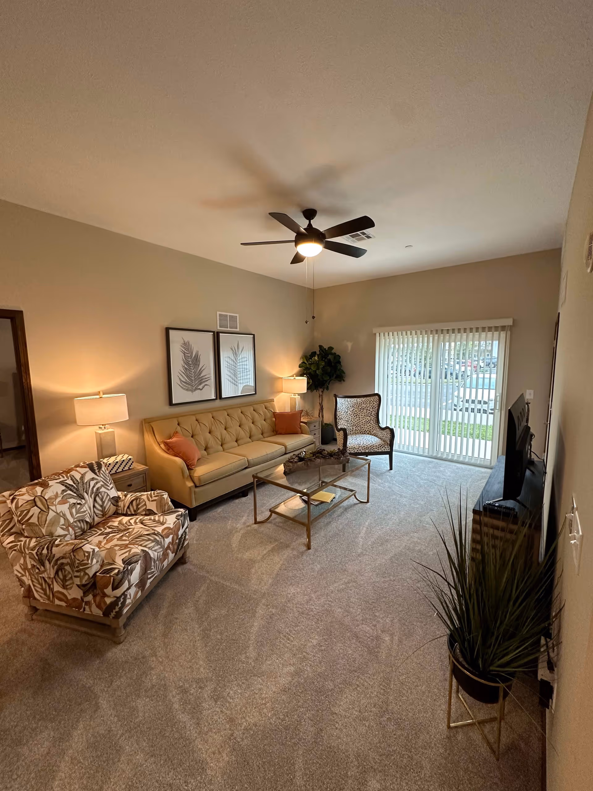 A cozy living room with a beige tufted sofa, two patterned armchairs, a glass coffee table, two side tables with lamps, a ceiling fan with light, a flat-screen TV on a stand, a potted plant, and vertical blinds covering a sliding glass door leading outside.
