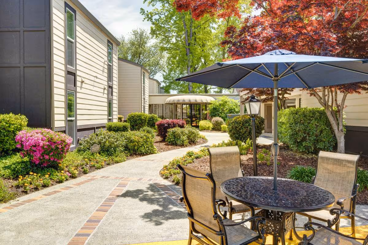 Outdoor patio area at Greenhaven Place featuring a round metal table with four chairs and a large blue umbrella. The patio is surrounded by well-maintained bushes, flowering plants, and trees with red and green leaves. Beige buildings with multiple windows border the walkway that curves through the garden area.