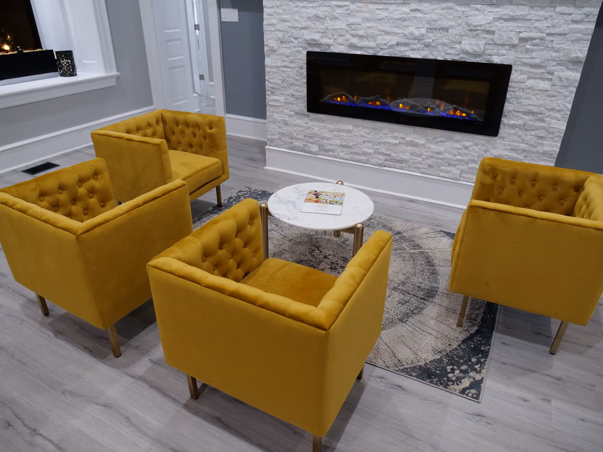 A cozy seating area with four mustard yellow armchairs arranged around a small round white marble table on a patterned rug. Behind the seating area is a modern electric fireplace set into a white textured stone wall. The room has light gray wood flooring and gray walls with white trim.