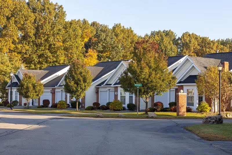 Row of single-story residential units with garages, landscaped trees, and a street in front under a clear sky.