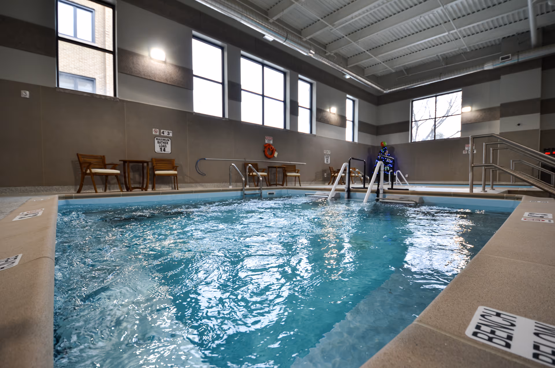 Indoor swimming pool with clear blue water, surrounded by beige tiled edges. There are several wooden chairs and small tables along the wall, with large windows letting in natural light. Handrails lead into the pool, and a person wearing a blue outfit is standing near the poolside. The ceiling is high with exposed beams and lighting fixtures.