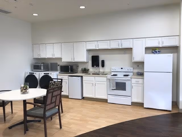 A clean and modern kitchen area with white cabinets, a white refrigerator, a white stove with an oven, a dishwasher, and two microwaves on the countertop. There is a round table with four chairs in the foreground, and a washing machine and dryer are visible on the left side. The floor has a combination of light wood and dark wood finishes.