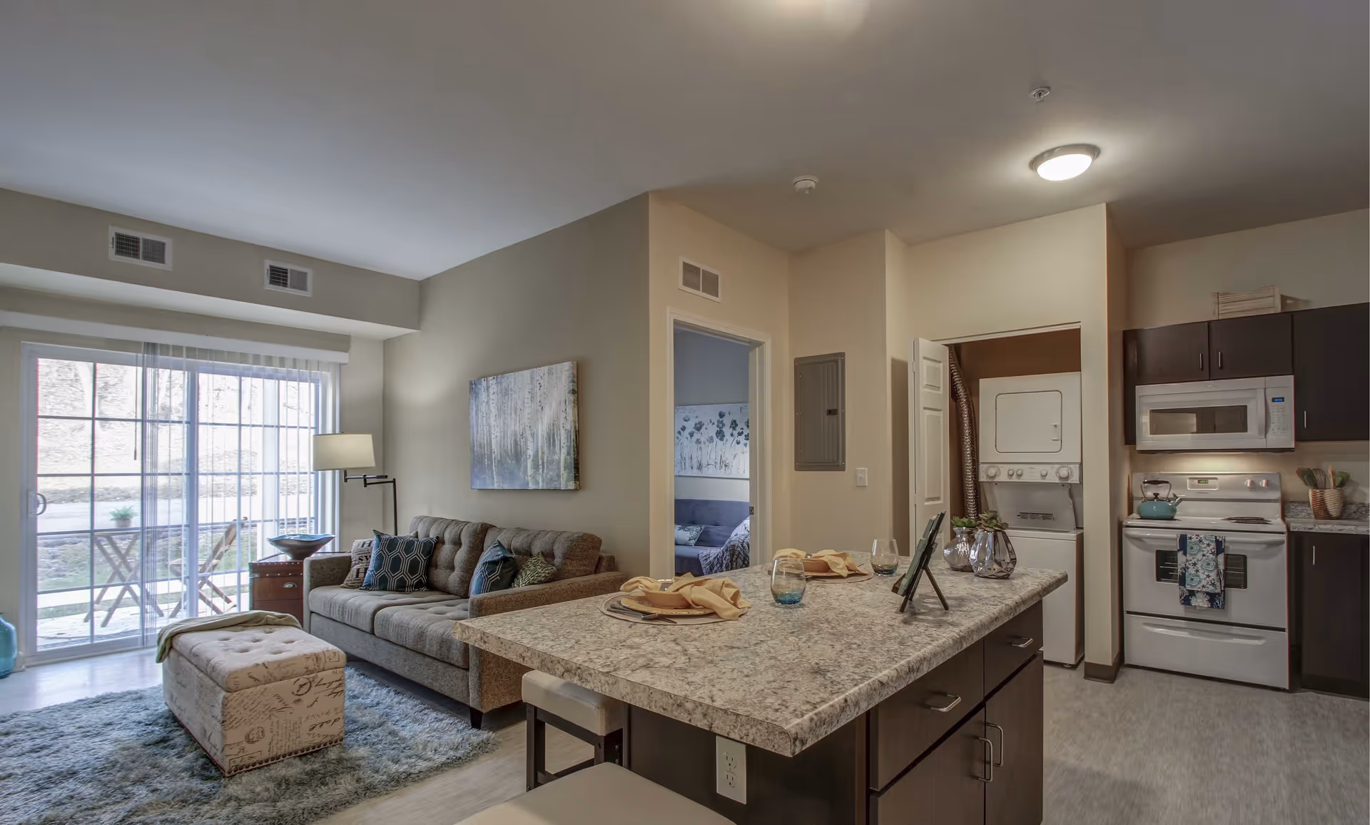 Interior view of a modern apartment featuring a kitchen with dark cabinets, a white stove, microwave, and a stacked washer and dryer. Adjacent to the kitchen is a living area with a gray sofa, a patterned ottoman, a floor lamp, and a large window with sliding glass doors leading outside. A dining counter with place settings and stools separates the kitchen and living area. A doorway leads to another room with a sofa and wall art.