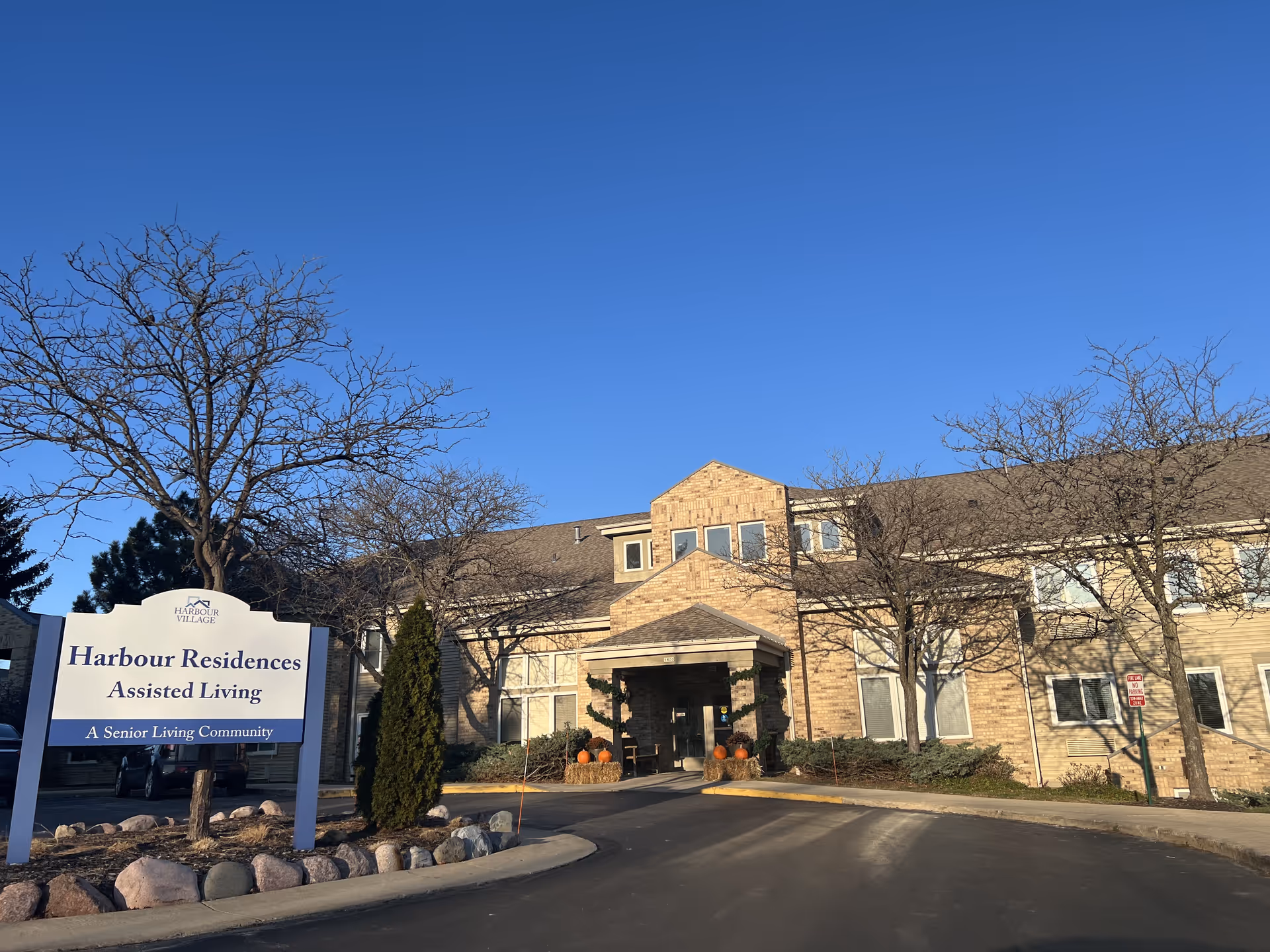Exterior view of Harbour Residences Assisted Living building with a clear blue sky. The building is two stories with a brick and siding facade. There are leafless trees and some landscaping around the entrance. A sign in front reads 'Harbour Residences Assisted Living A Senior Living Community.'