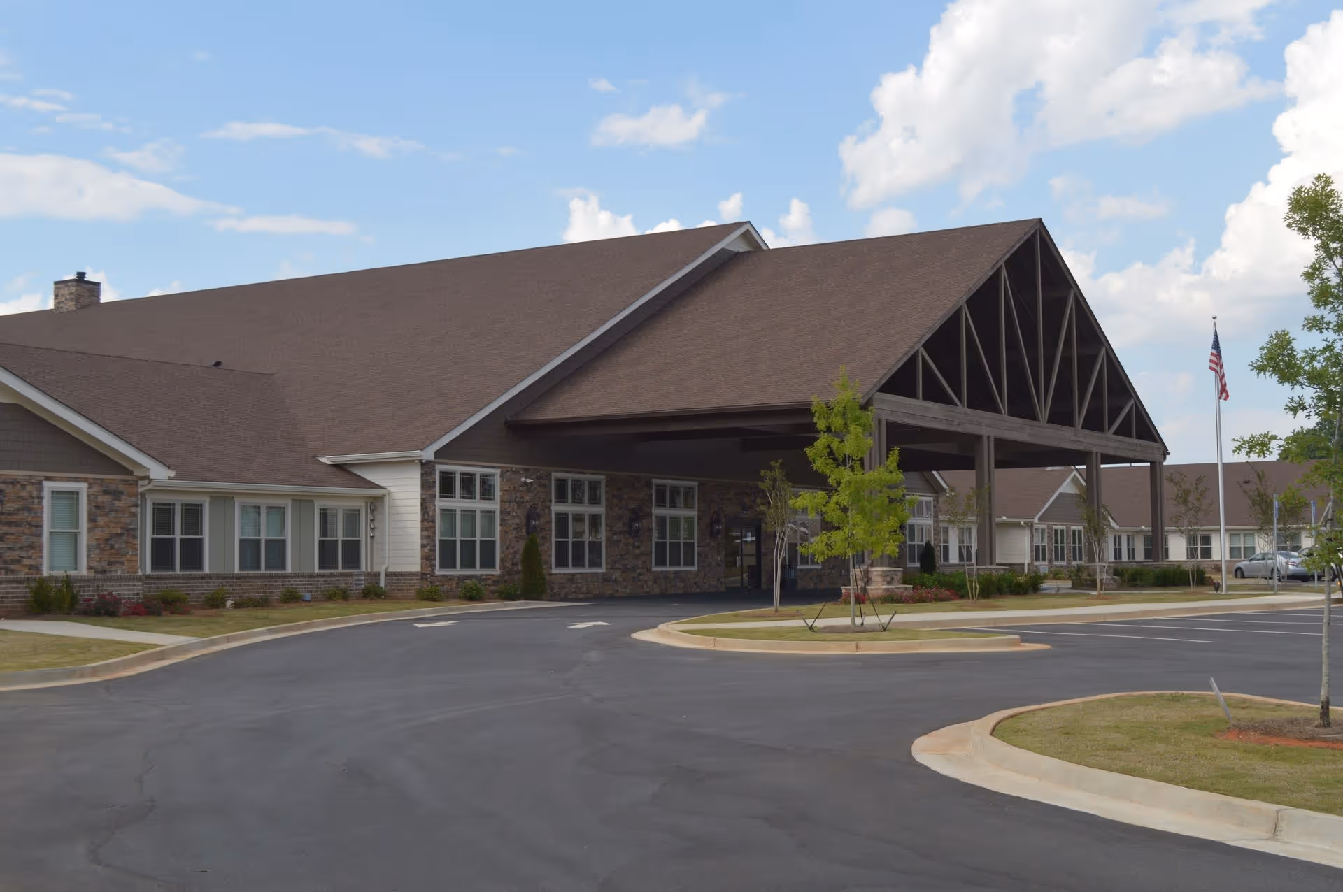 Exterior view of The Phoenix at Lake Joy facility showing a large building with a covered entrance, stone and siding facade, multiple windows, a paved driveway, landscaped areas with small trees, and an American flag on a flagpole under a partly cloudy sky.