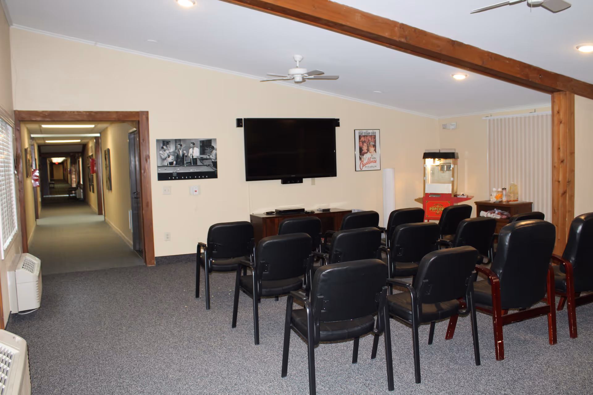 A small theater or media room with several rows of black chairs facing a wall-mounted flat screen TV. There are two framed movie posters on the wall, a popcorn machine in the corner, and a long hallway visible to the left. The room has beige walls, a carpeted floor, and wooden beams on the ceiling.