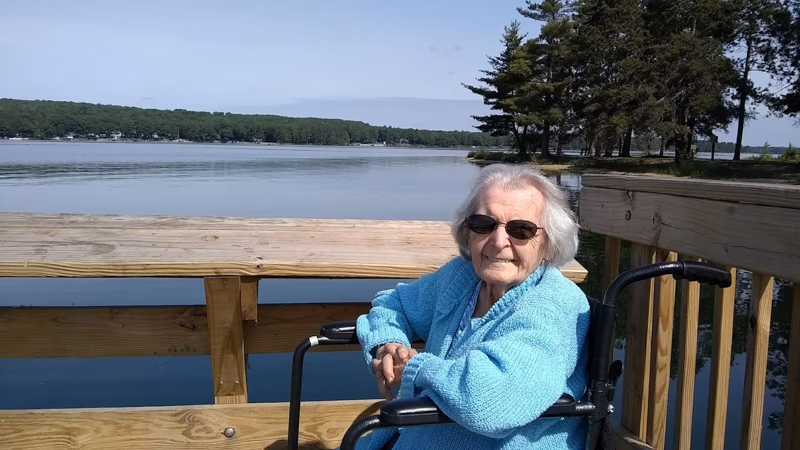 An elderly woman wearing sunglasses and a blue sweater sits in a wheelchair on a wooden dock by a calm lake with trees in the background under a clear sky.