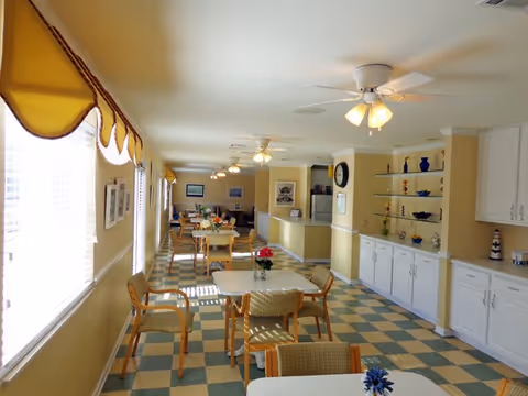 A bright dining area with multiple tables and chairs arranged neatly. The room has large windows with yellow valances allowing natural light to fill the space. The floor features a checkered pattern in green and beige. Ceiling fans with lights are mounted on the ceiling. On the right side, there are white cabinets and shelves displaying decorative items.