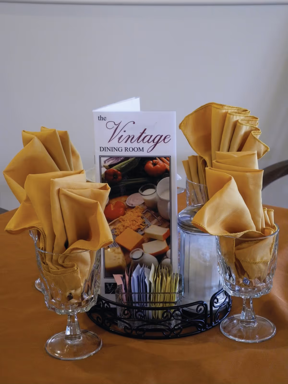 A table setting with four crystal glasses each holding a neatly folded yellow cloth napkin. In the center, there is a menu card titled 'the Vintage DINING ROOM' featuring images of cheese, vegetables, and milk. The tablecloth is yellowish-orange.