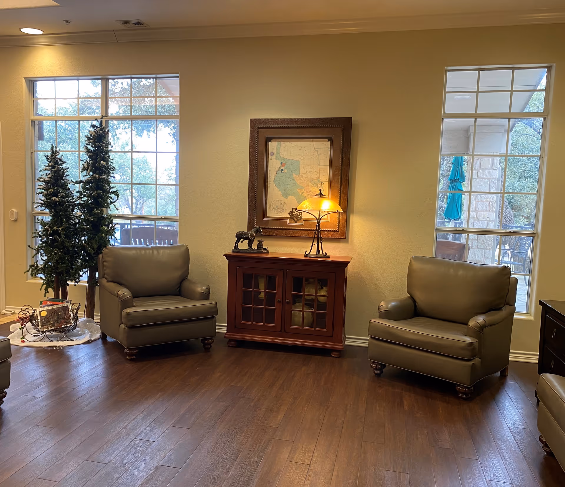 A cozy sitting area with two green armchairs facing each other, a wooden cabinet with a lit table lamp and a small horse figurine on top, a framed map on the wall, and two large windows showing outdoor trees and patio furniture.