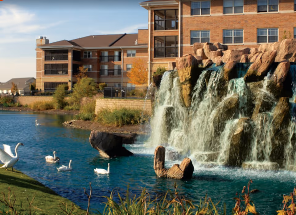 A scenic outdoor view of Alden of Waterford featuring a large artificial waterfall cascading into a pond with swans swimming. In the background, there is a multi-story brick building with multiple windows and balconies. The area is surrounded by greenery and landscaping.