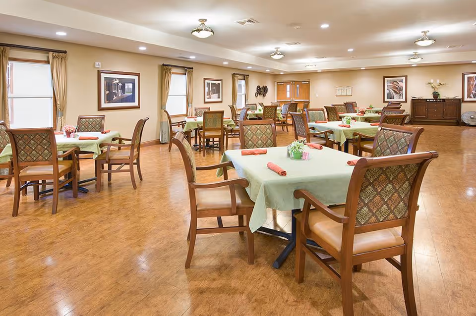A spacious dining room with multiple tables covered in light green tablecloths, each set with a small flower arrangement and rolled red napkins. The room has wooden chairs with patterned backs and cushioned seats, wood flooring, beige walls, framed artwork, and windows with curtains. Ceiling lights illuminate the room.