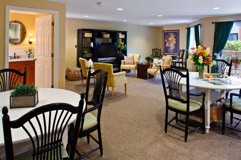A cozy common area in a senior living facility featuring multiple seating arrangements including cushioned armchairs and black wooden chairs around white tables. One table has a puzzle and a vase with colorful flowers. The room has beige walls, carpeted floors, and large windows with green curtains letting in natural light. A black entertainment center with a TV and decorative items is against one wall. To the left, an open door reveals a bathroom with a round mirror and a sink.