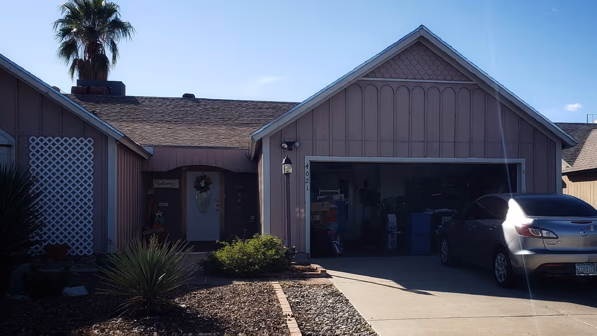 Front exterior view of a single-story residential building with a peaked roof, a garage with an open door showing stored items inside, a driveway with a parked silver car, some landscaping with bushes and a palm tree, and a front door decorated with a wreath.
