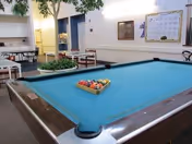 Interior view of a senior living facility common area featuring a pool table with a rack of billiard balls set up for a game. In the background, there are tables and chairs, a kitchenette area, and framed artwork on the walls. The space is well-lit and decorated with plants.