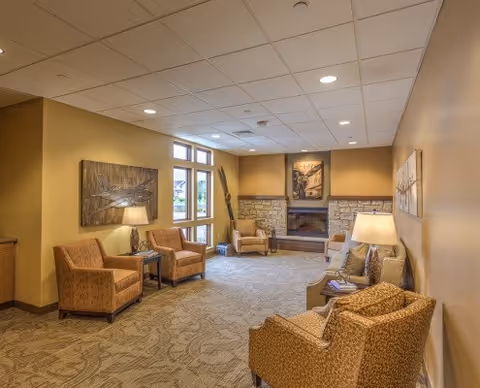 A cozy living room area in an assisted living facility featuring several upholstered armchairs arranged around a stone fireplace. The walls are painted a warm beige color and decorated with framed artwork. There are table lamps providing soft lighting, and large windows letting in natural light.