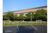 Exterior view of a brick building with large windows partially obscured by trees and greenery. In the foreground, there is a parking lot with marked spaces including a handicapped parking spot. A wooden fence runs parallel to the building.