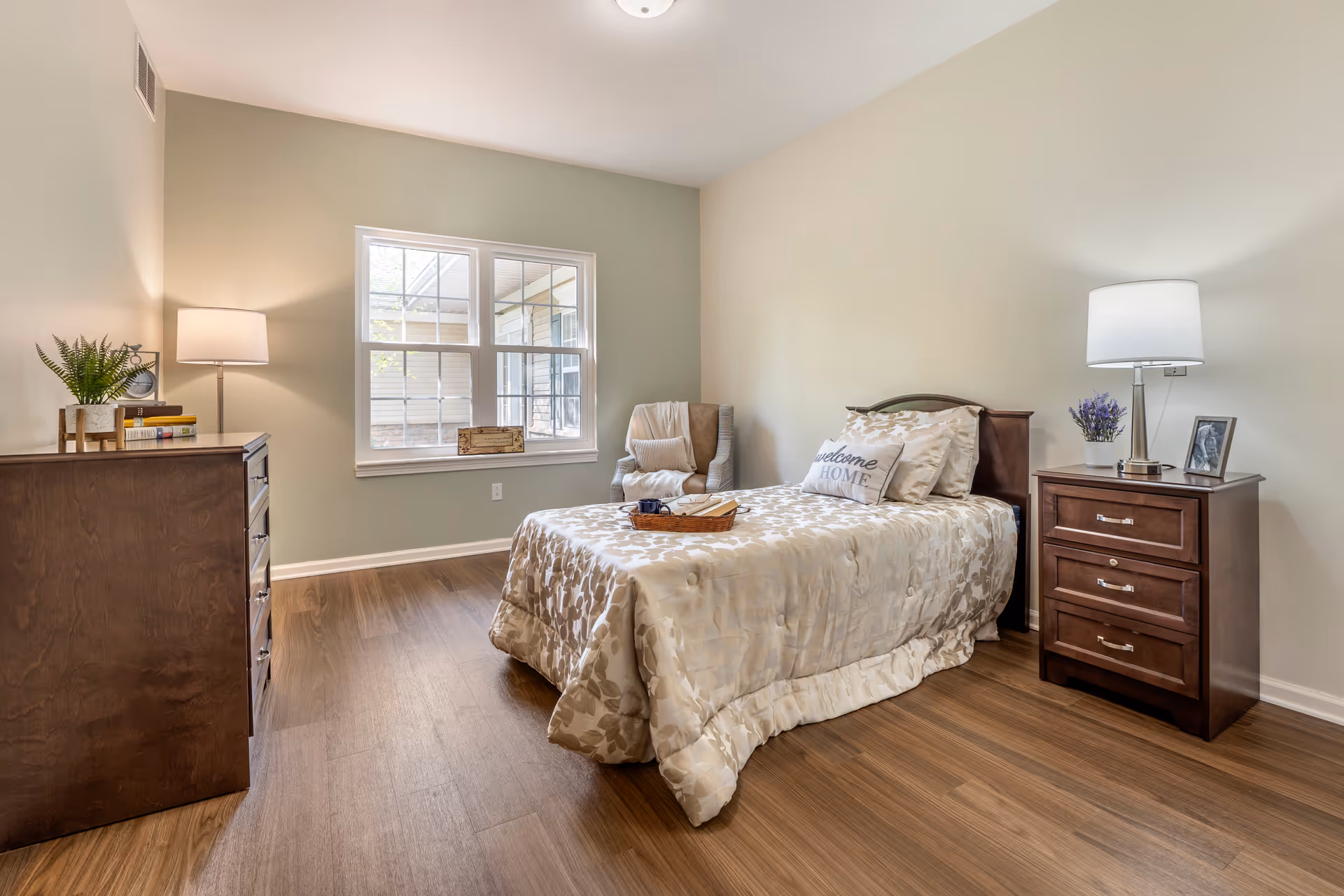 A neatly arranged bedroom with a single bed covered in a beige floral comforter and a 'welcome HOME' pillow. There is a wooden nightstand with a lamp, a small plant, and a framed photo on it. Across from the bed is a wooden dresser with a lamp, books, and a small plant. A cushioned armchair with a throw blanket and pillow is placed near a window that lets in natural light. The room has wooden flooring and light-colored walls.