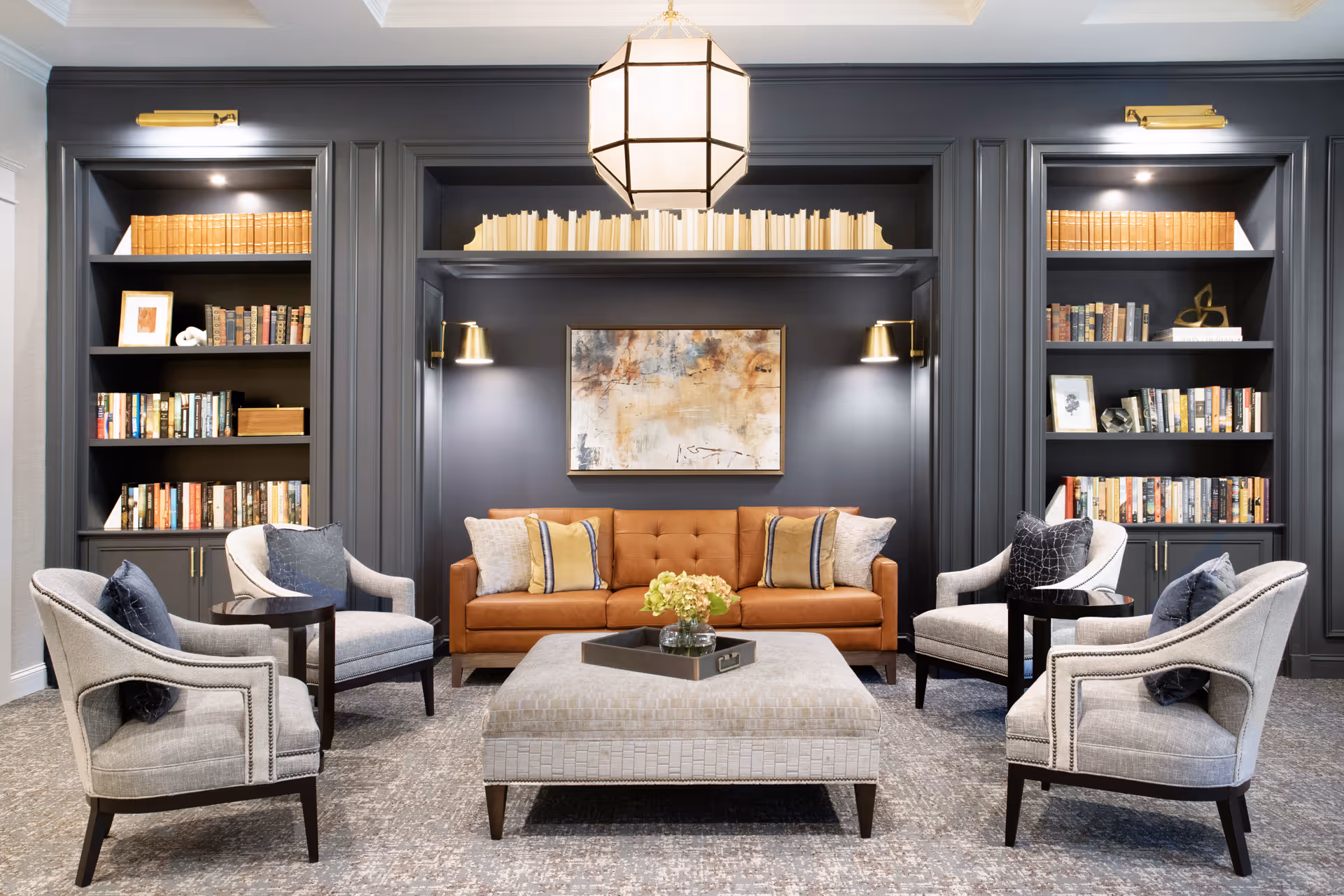 Comfortable lounge with a brown leather sofa, four upholstered armchairs around a central ottoman, and built-in bookcases and artwork on a dark paneled wall.