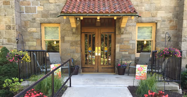Entrance to a senior living facility with double wooden doors under a red tiled awning, flanked by two windows. There are black metal railings on either side of the concrete walkway leading to the doors, with hanging flower baskets and potted plants adding color to the scene.