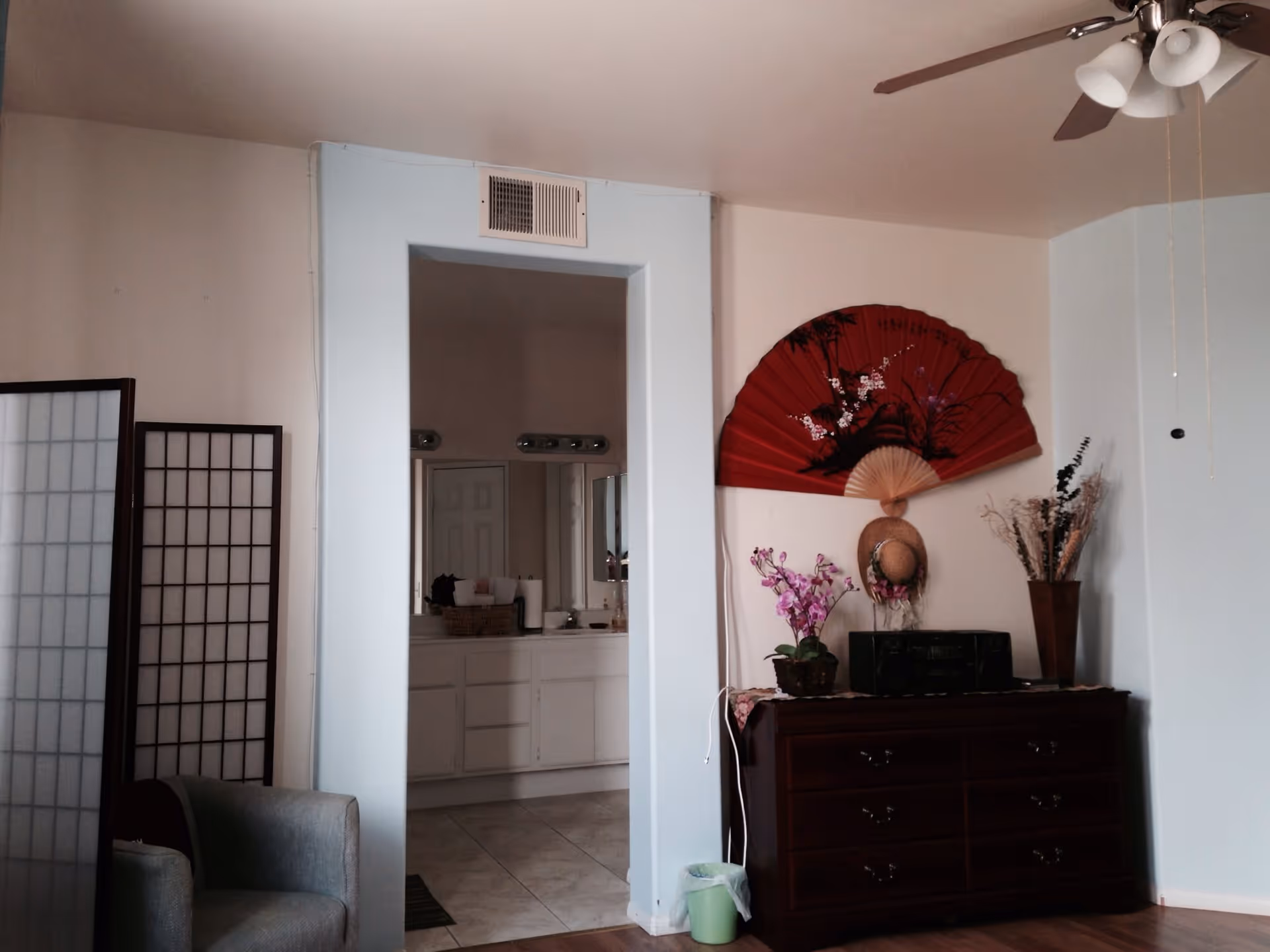 Interior view of a senior living facility room showing a gray armchair next to a folding screen on the left, an open doorway leading to a bathroom with a sink and mirror, and a dark wooden dresser on the right with decorative items including a large red fan, a hat, a pink orchid, and a vase with dried plants. A ceiling fan with lights is visible above.