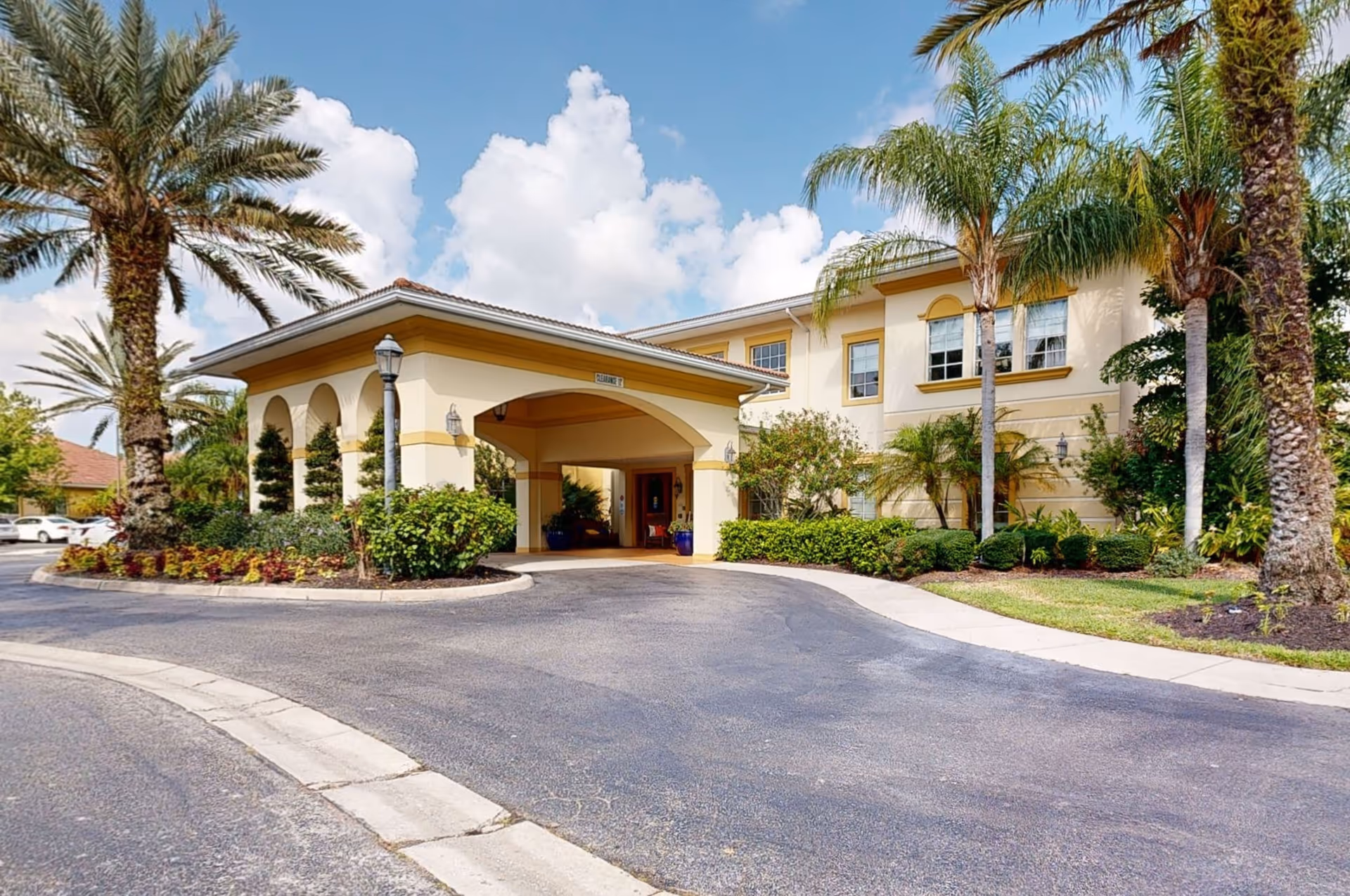 Exterior view of The Windsor of Lakewood Ranch facility entrance with a covered drop-off area, surrounded by palm trees and landscaped greenery under a partly cloudy sky.