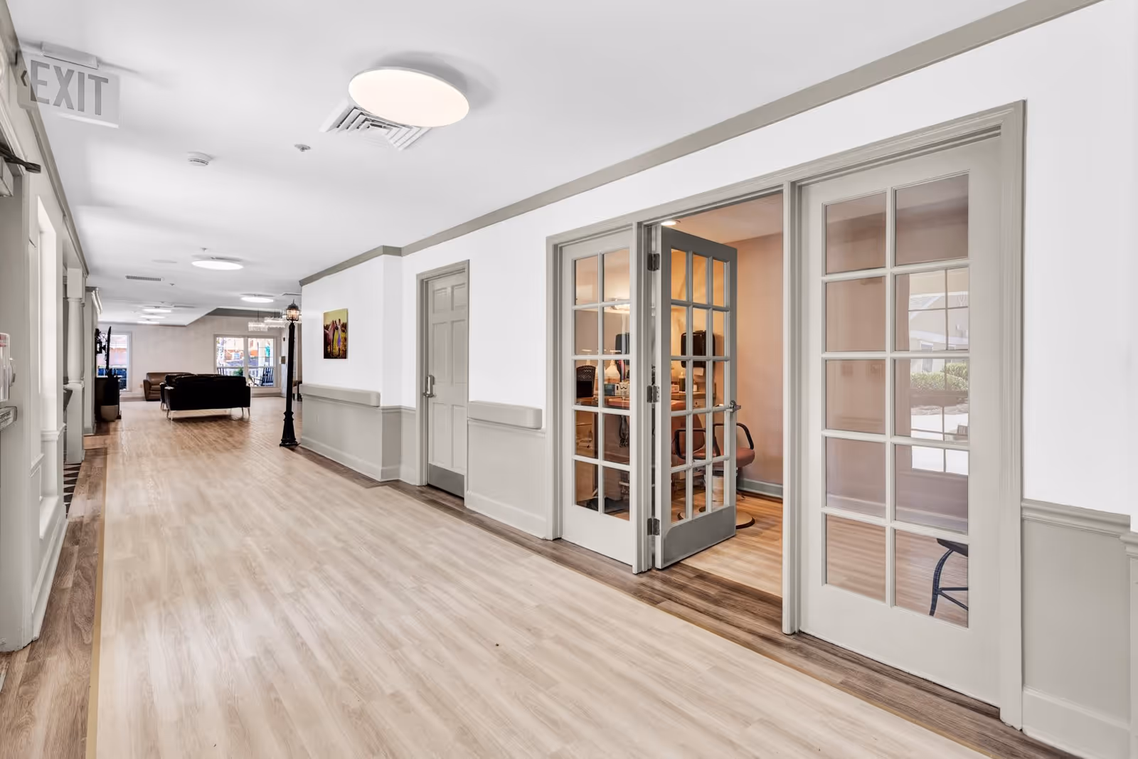 A bright and clean hallway in an assisted living facility with light wood flooring and white walls with gray trim. On the right side, there are glass-paneled double doors leading to a room with office furniture. At the end of the hallway, there is a seating area with sofas and large windows letting in natural light. An exit sign is visible on the left side near the ceiling.