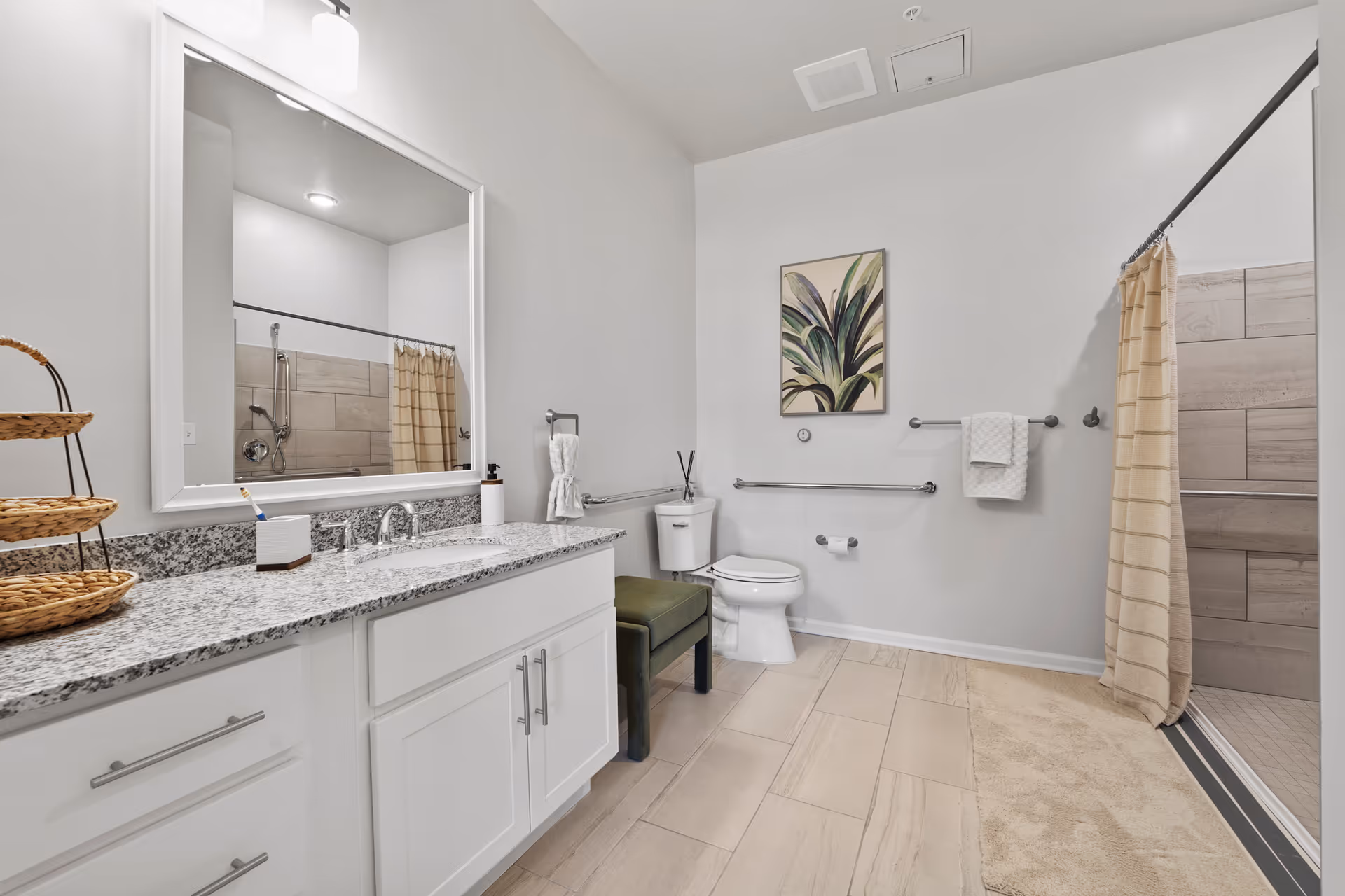 A clean and modern bathroom with a large mirror above a granite countertop with a sink. There is a white toilet next to a green cushioned bench. The shower area has beige tiles and a beige shower curtain. The walls are white, and there is a framed plant artwork hanging above the toilet. The floor is tiled in a light beige color, and there are grab bars installed near the toilet and shower area.