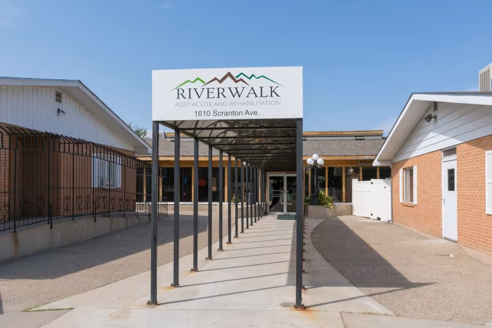 Entrance walkway to Riverwalk Post Acute and Rehabilitation facility at 1610 Scranton Ave, featuring a covered metal frame pathway leading to the building entrance with brick walls and a clear blue sky.