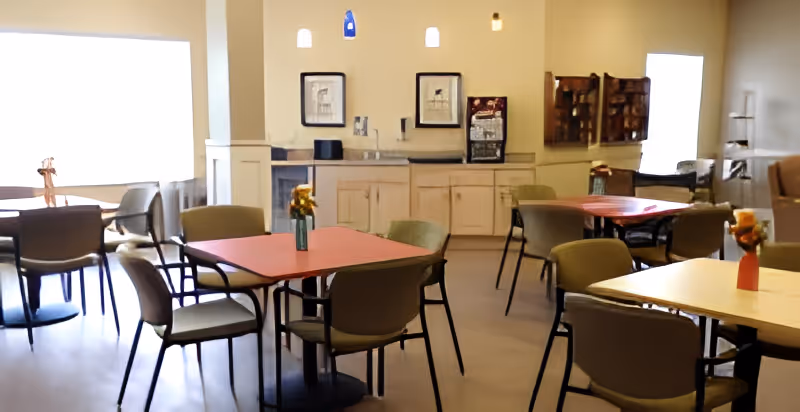 Dining area with several tables and chairs, small vases of flowers on the tables, and a counter with cabinets and a beverage station.