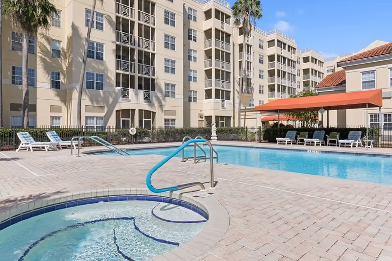 Outdoor swimming pool area with a hot tub in the foreground, surrounded by lounge chairs and a building with multiple floors and balconies in the background under a clear blue sky.