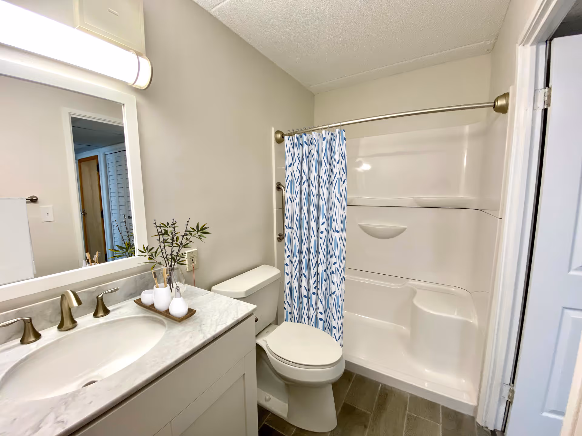 A clean and modern bathroom featuring a white toilet, a white bathtub with a built-in seat and a blue and white patterned shower curtain. There is a white vanity with a marble countertop, a sink with brushed nickel faucets, a large mirror, and a light fixture above the mirror. A small tray with toothbrushes and a vase with green foliage is placed on the countertop. The floor has gray tiles and the walls are painted light beige.