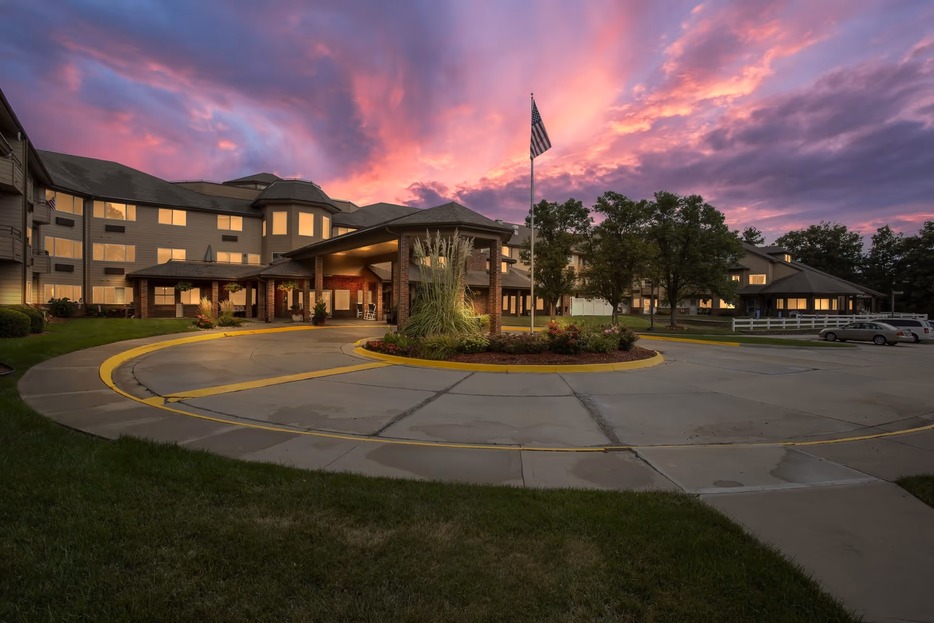 Exterior view of Holiday Country Squire senior living facility at sunset with a dramatic pink and purple sky, a circular driveway with landscaping and an American flag in front of the building.