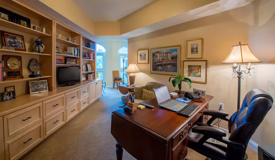 A cozy office space with a wooden desk holding a laptop, keyboard, and office supplies. Behind the desk is a comfortable leather office chair. To the left, there is a large built-in wooden bookshelf filled with books, framed photos, and decorative items. The room is warmly lit by a floor lamp and a table lamp, with framed artwork hanging on the beige walls. In the background, there is a small seating area near windows letting in natural light.
