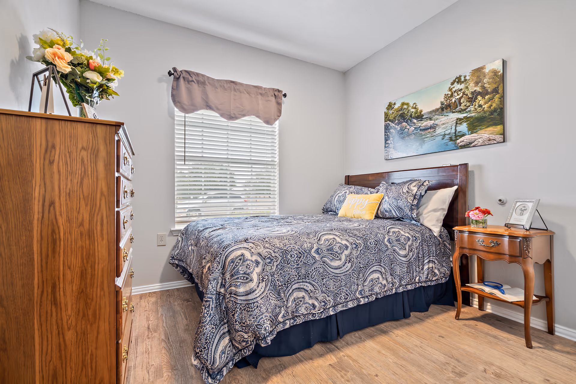 Neatly furnished bedroom with a patterned bed, wooden dresser and nightstand, window with blinds, and landscape artwork above the bed.