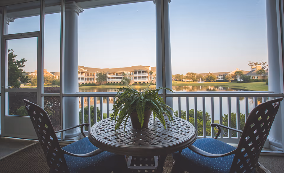 A screened-in porch with a round wooden table and two blue cushioned chairs. A potted fern sits on the table. Beyond the porch, there is a view of a pond with reflections of nearby buildings and trees under a clear sky.