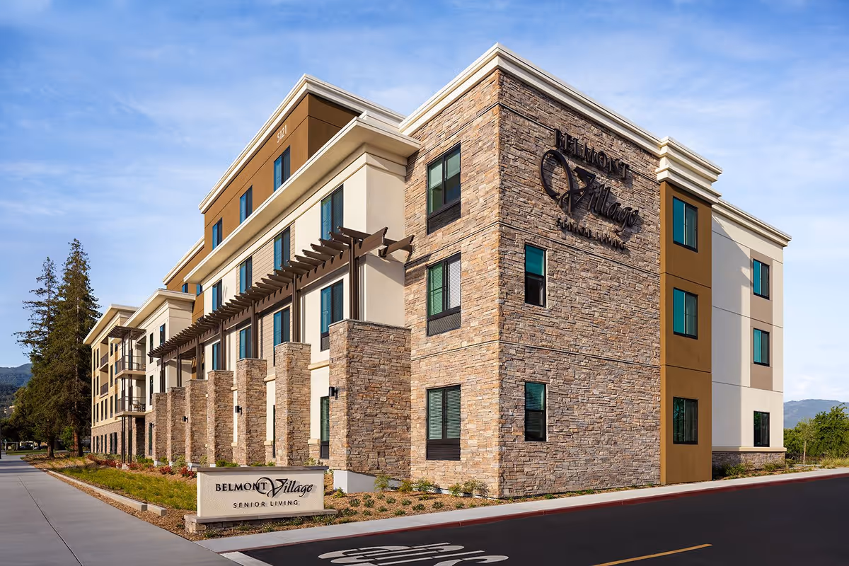 Exterior view of Belmont Village Senior Living Los Gatos building with stone and beige facade, multiple windows, and a sign in front displaying the facility name.