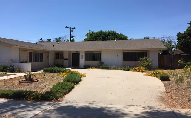 Single-story white building with a gray shingled roof, featuring several windows with bars and a central entrance door. The front yard has a concrete driveway with some green shrubs and yellow flowers along the edges. Trees and a utility pole are visible in the background under a clear blue sky.