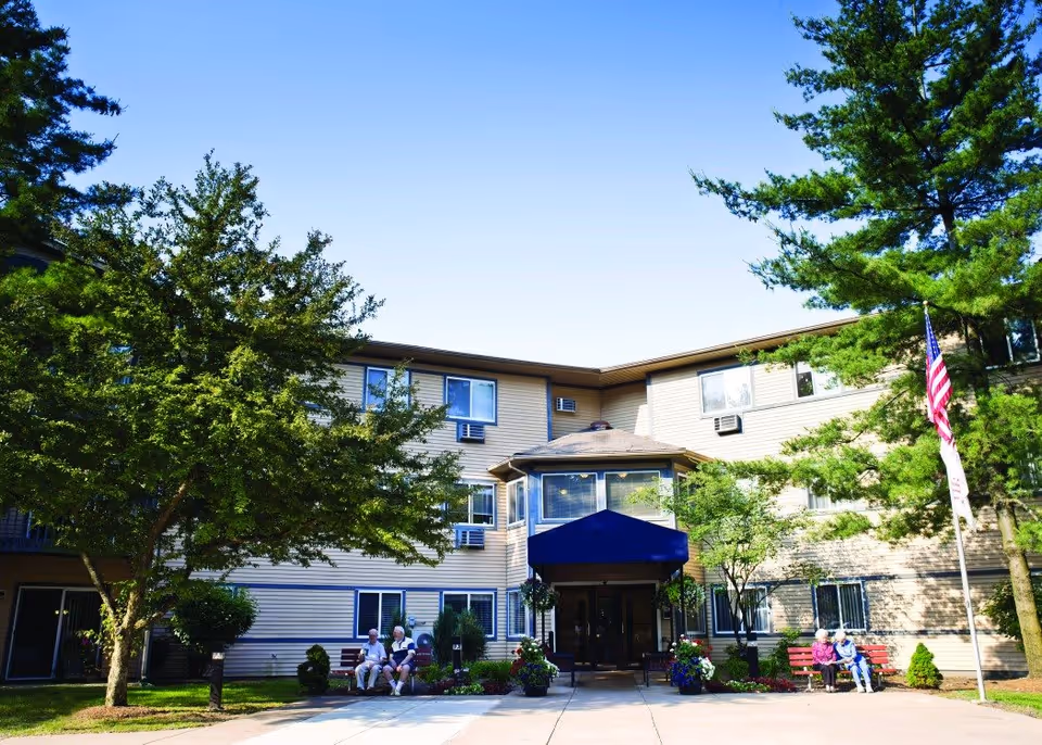 Exterior view of a three-story senior living facility with beige siding and multiple windows. There is a blue awning over the main entrance, surrounded by greenery including trees and shrubs. Several elderly people are sitting on benches near the entrance. An American flag is displayed on a flagpole to the right.