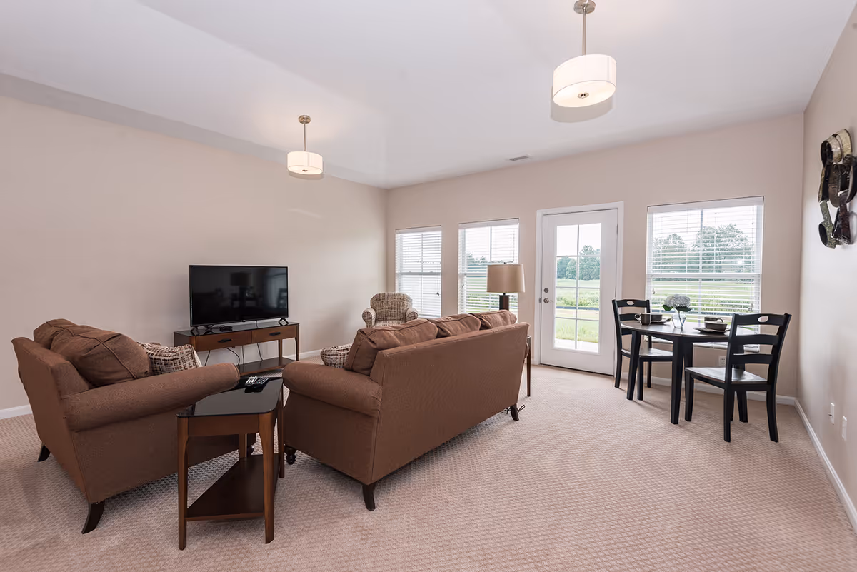 Bright living room with brown sofas facing a TV, a small dining table by windows and a glass door to the outside.