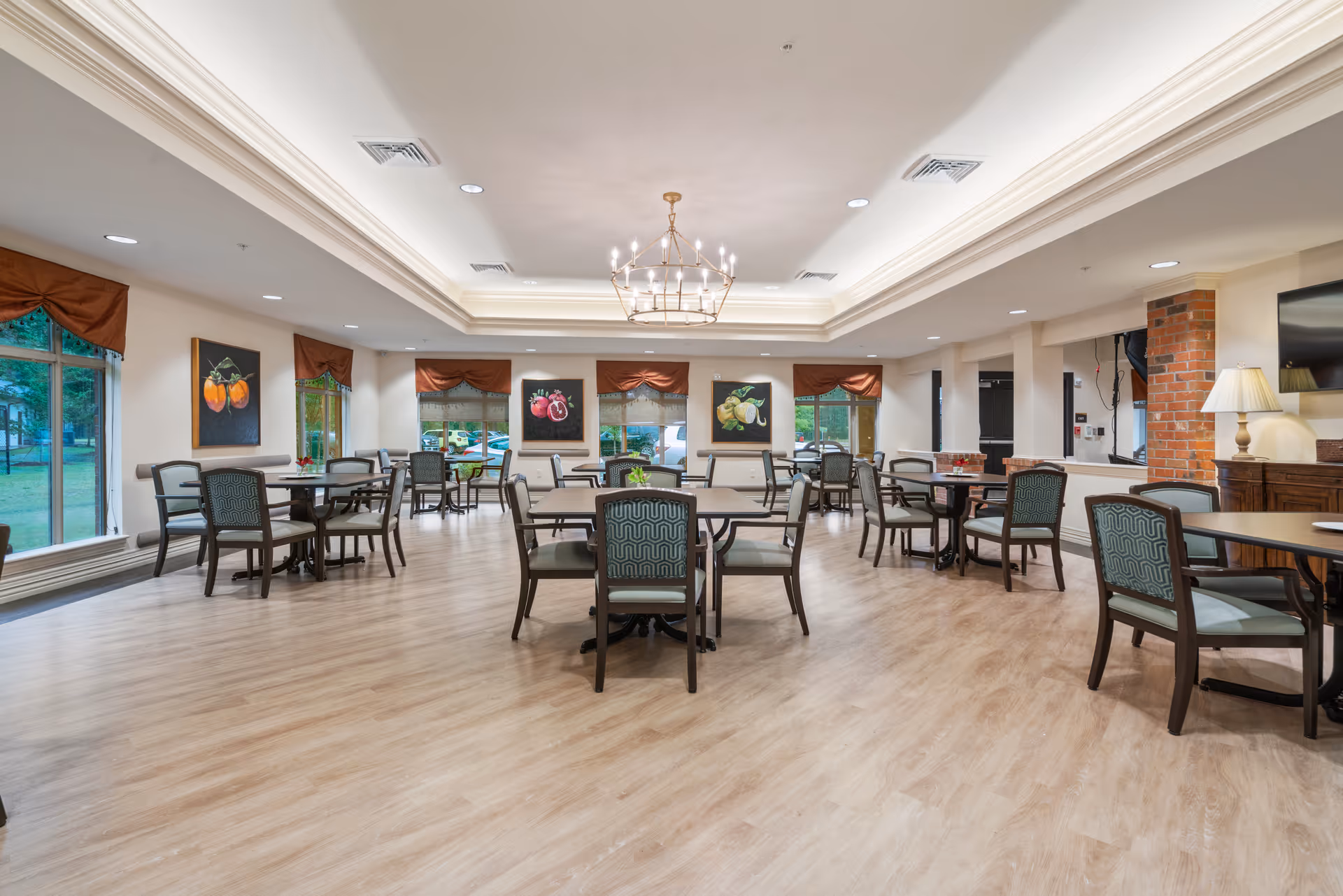 A spacious dining room with multiple tables and chairs arranged neatly. The room has large windows with brown valances, light wood flooring, and a chandelier hanging from a recessed ceiling. There are paintings of fruit on the walls and a lamp on a side table near a brick column.