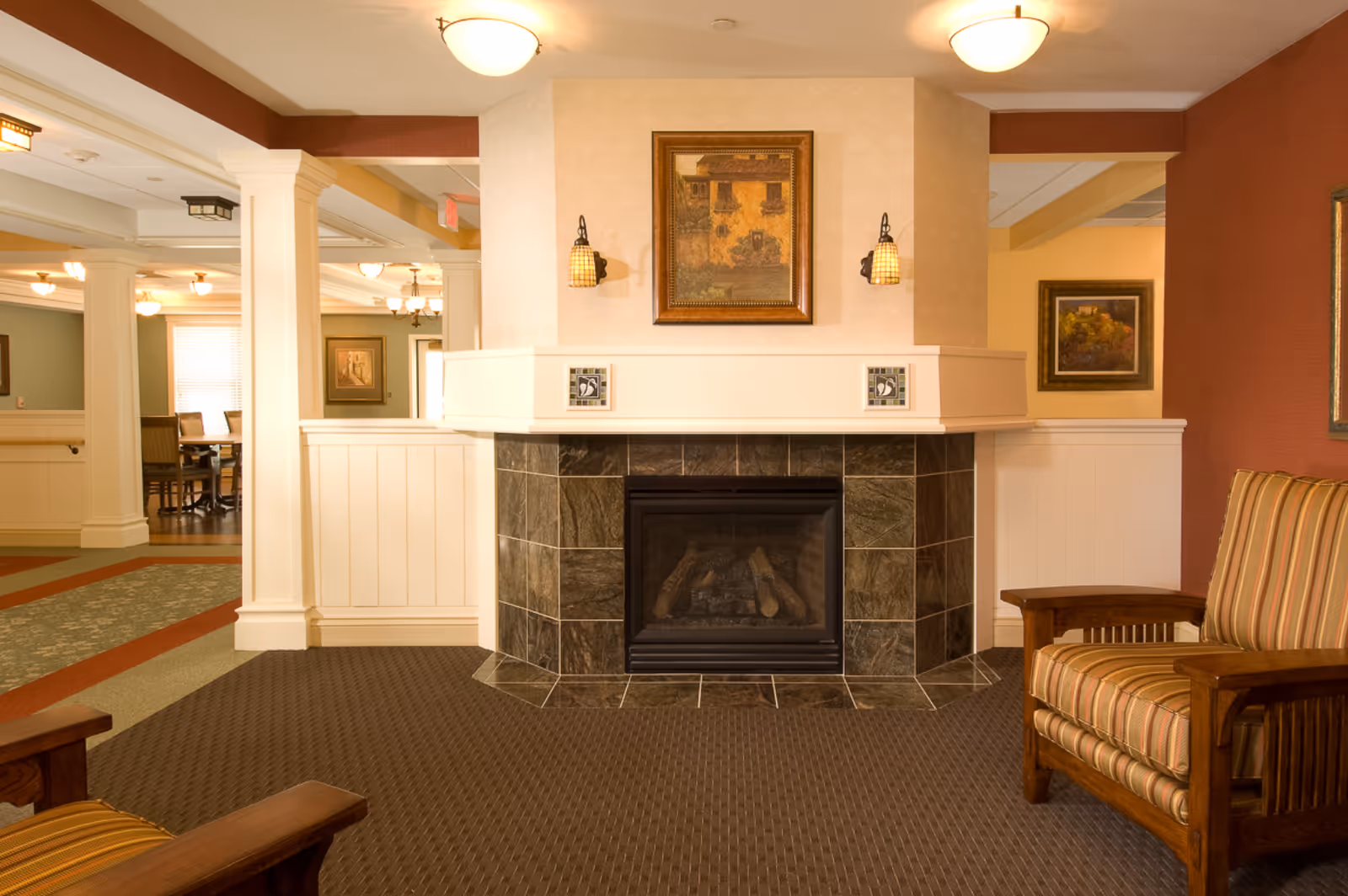 A cozy interior lounge area featuring a tiled fireplace with a framed painting above it, flanked by two wall sconces. The room has warm lighting, patterned carpet, and wooden armchairs with striped cushions. In the background, there is a dining area with tables and chairs, separated by white columns and half walls.