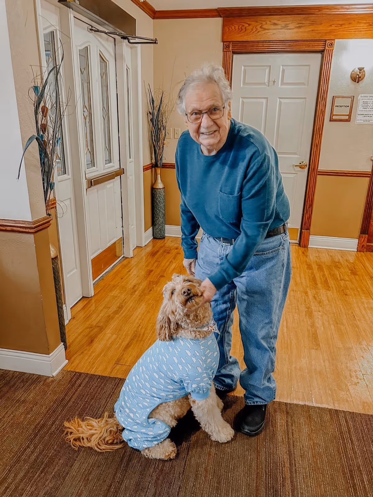 An elderly man wearing glasses, a teal long-sleeve shirt, and jeans is standing indoors on a wooden floor, smiling and petting a large dog dressed in a blue outfit with white patterns. The background shows a door, a reception sign, and decorative vases with tall plants.