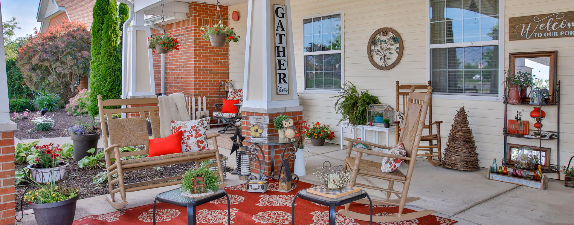 A cozy outdoor porch area with wooden rocking chairs and a bench adorned with colorful cushions and pillows. The porch features a red patterned rug, various potted plants, decorative items including a 'GATHER here' sign on a column, and a shelving unit with a 'Welcome to our porch' sign. The background shows part of a brick building and a garden with greenery and flowers.