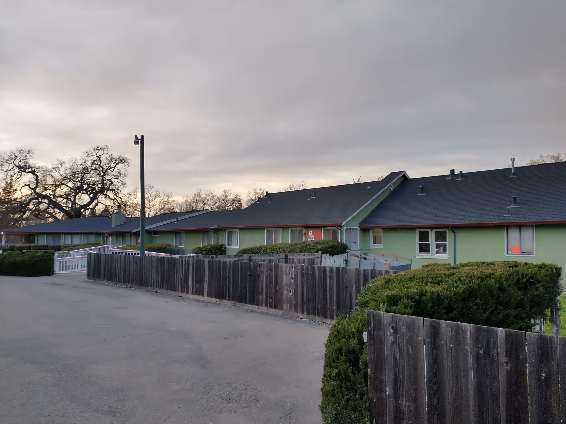 Exterior view of a single-story green building with a dark roof, surrounded by wooden fences and trimmed bushes under a cloudy sky.