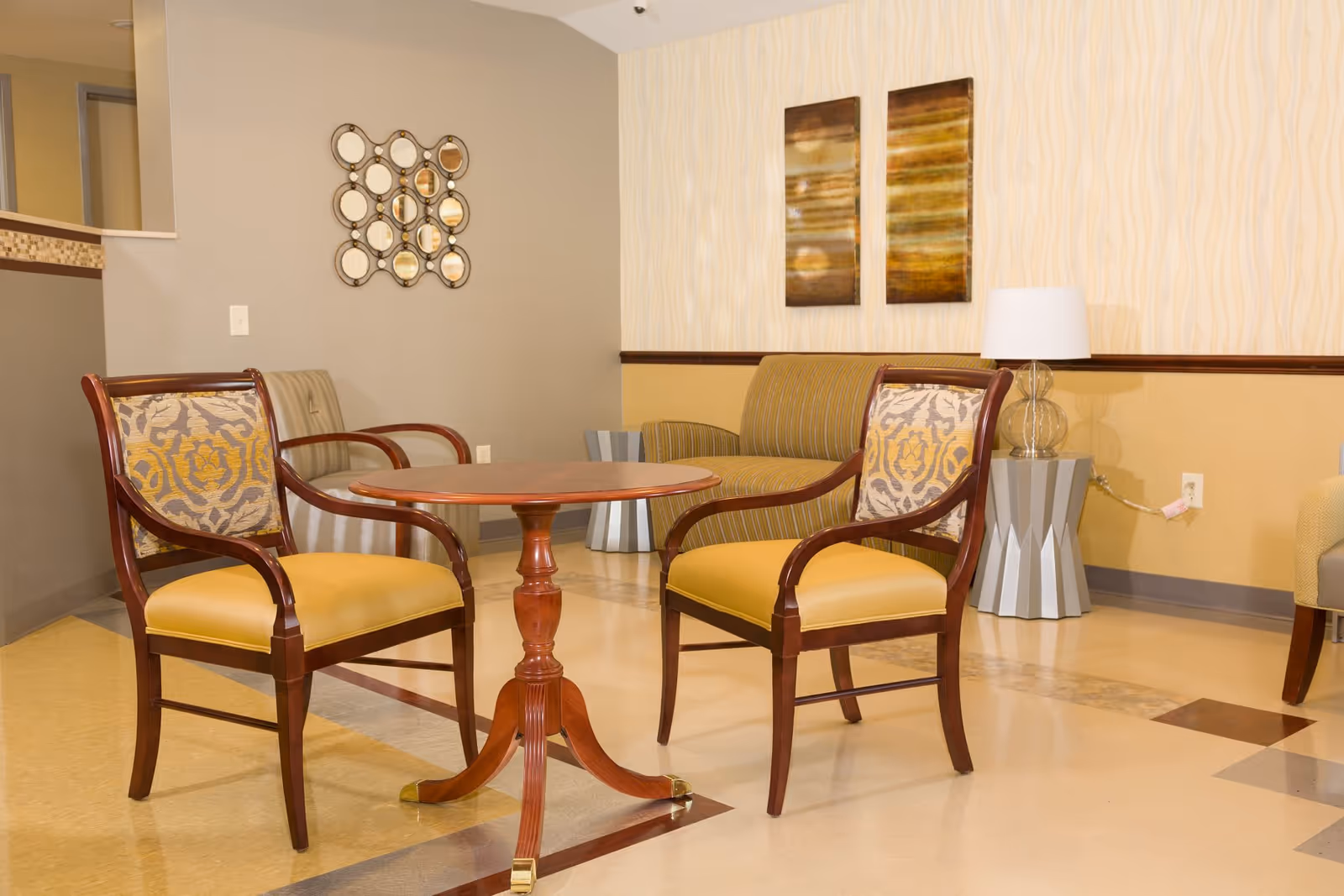 A furnished lounge area with two wooden armchairs around a small round table and a couch against a patterned wall.
