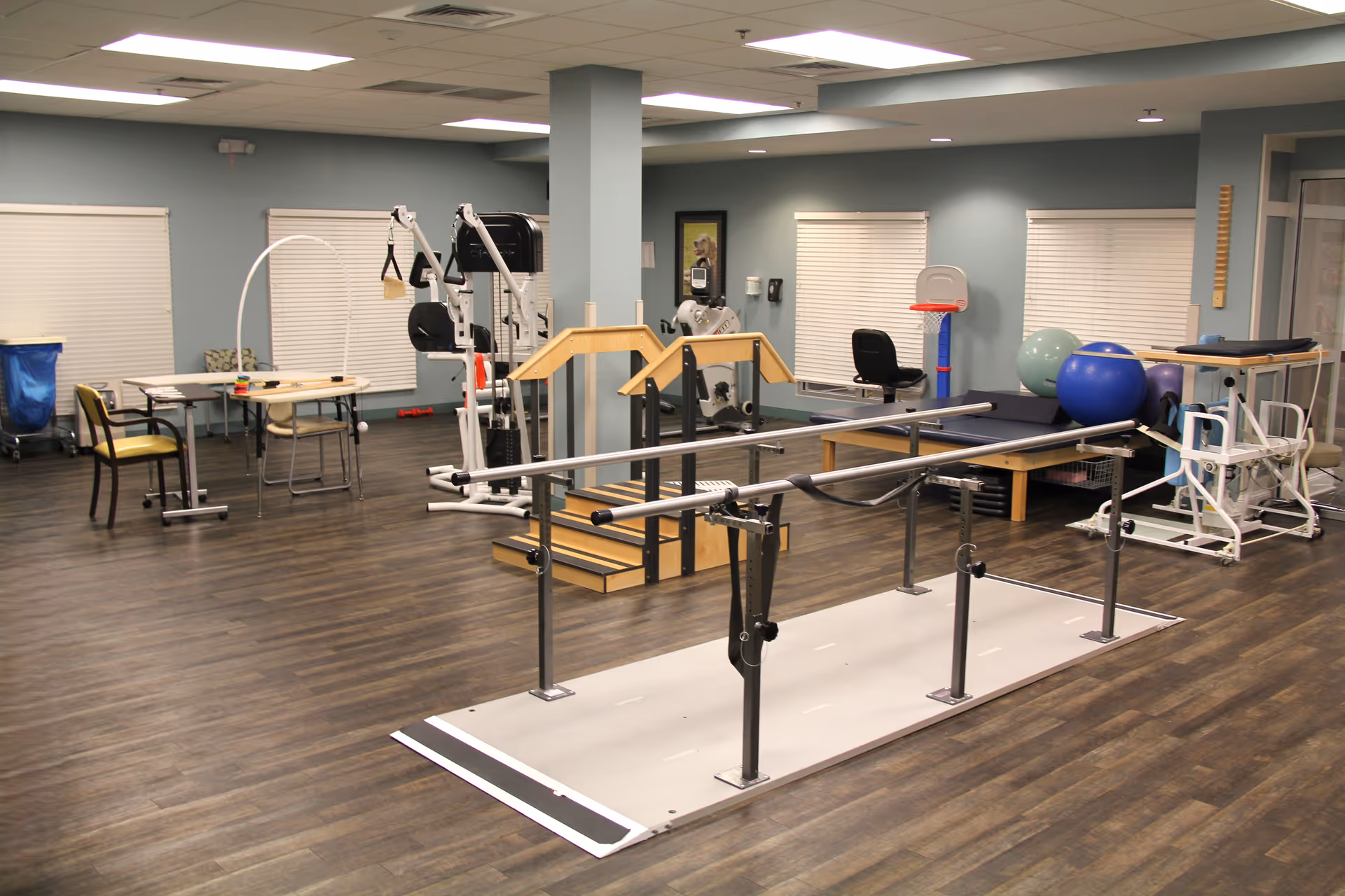 A spacious rehabilitation room with parallel bars, exercise equipment, therapy balls, and tables and chairs.